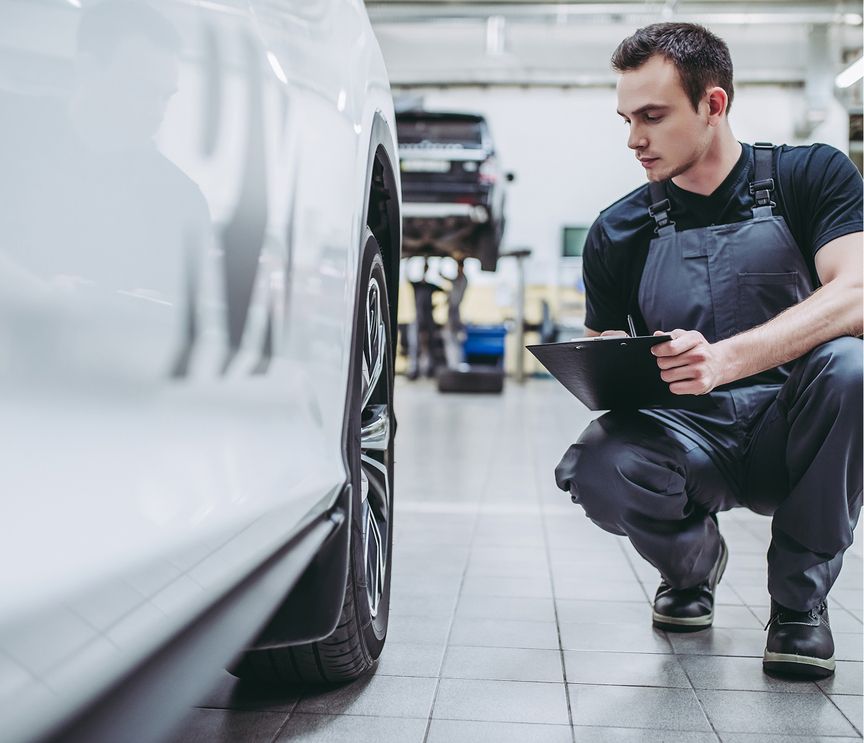 Mann in Arbeitskleidung kniet neben einem weißen Auto und hält ein Tablet in einer Werkstatt